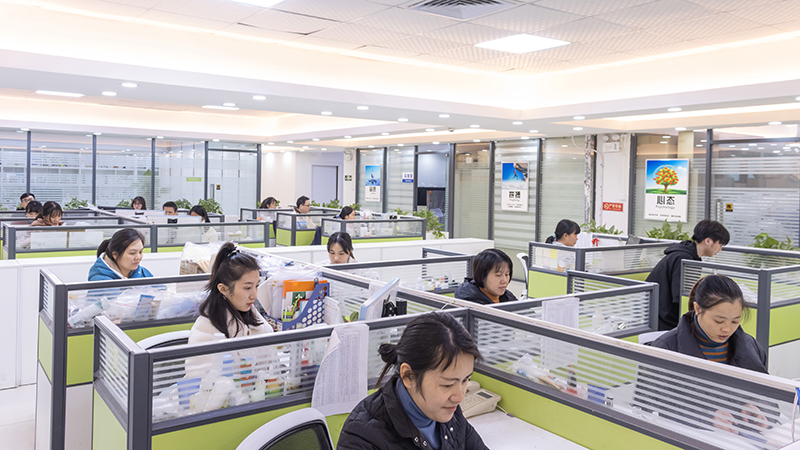 Office workspace: Teams manage eco - friendly, reusable glass cosmetic bottle business Office workers in cubicles, likely handling orders for eco - friendly glass cosmetic/dropper bottles